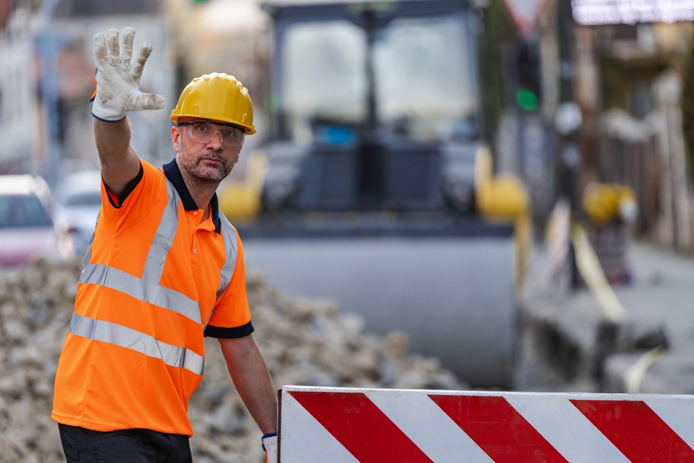 Construction worker in a hard hat and high-visibility vest raising his hand to signal traffic, standing at a roadwork site with heavy machinery and barriers in the background.