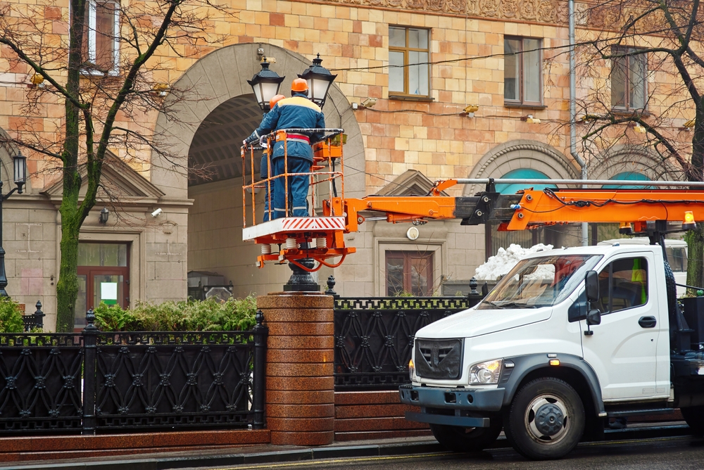 A worker in safety gear stands on an elevated cherry picker platform attached to a truck, repairing or maintaining a streetlamp outside a historic-style building.