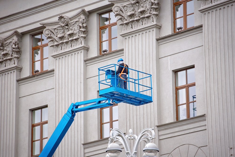A worker in a blue boom lift inspecting the facade of a historic building with classical columns, illustrating cherry picker hire for restoration projects. A worker in a blue boom lift inspecting the facade of a historic building with classical columns, illustrating cherry picker hire for restoration projects.