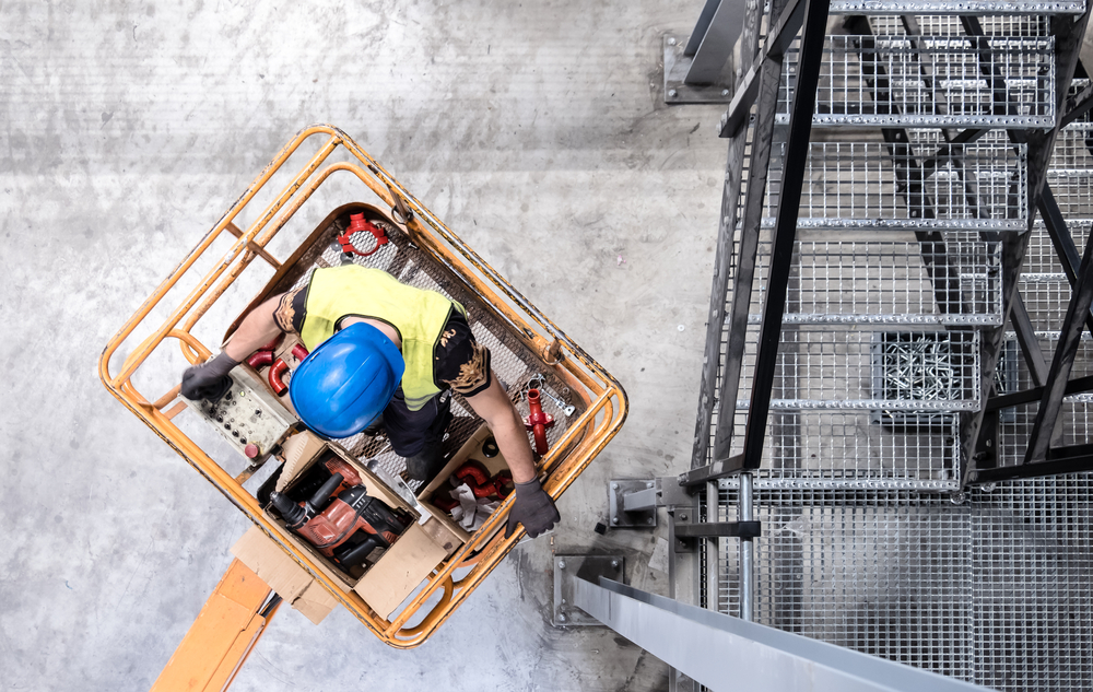 Overhead view of a worker with tools in an orange lift basket inside a warehouse, illustrating indoor cherry picker hire applications.
