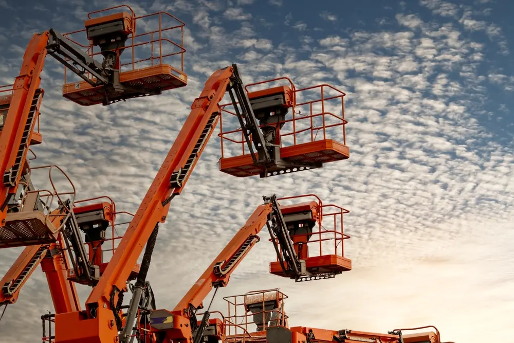 A fleet of orange articulated boom lifts with baskets raised against a cloudy sky, showcasing inventory available for cherry picker hire.